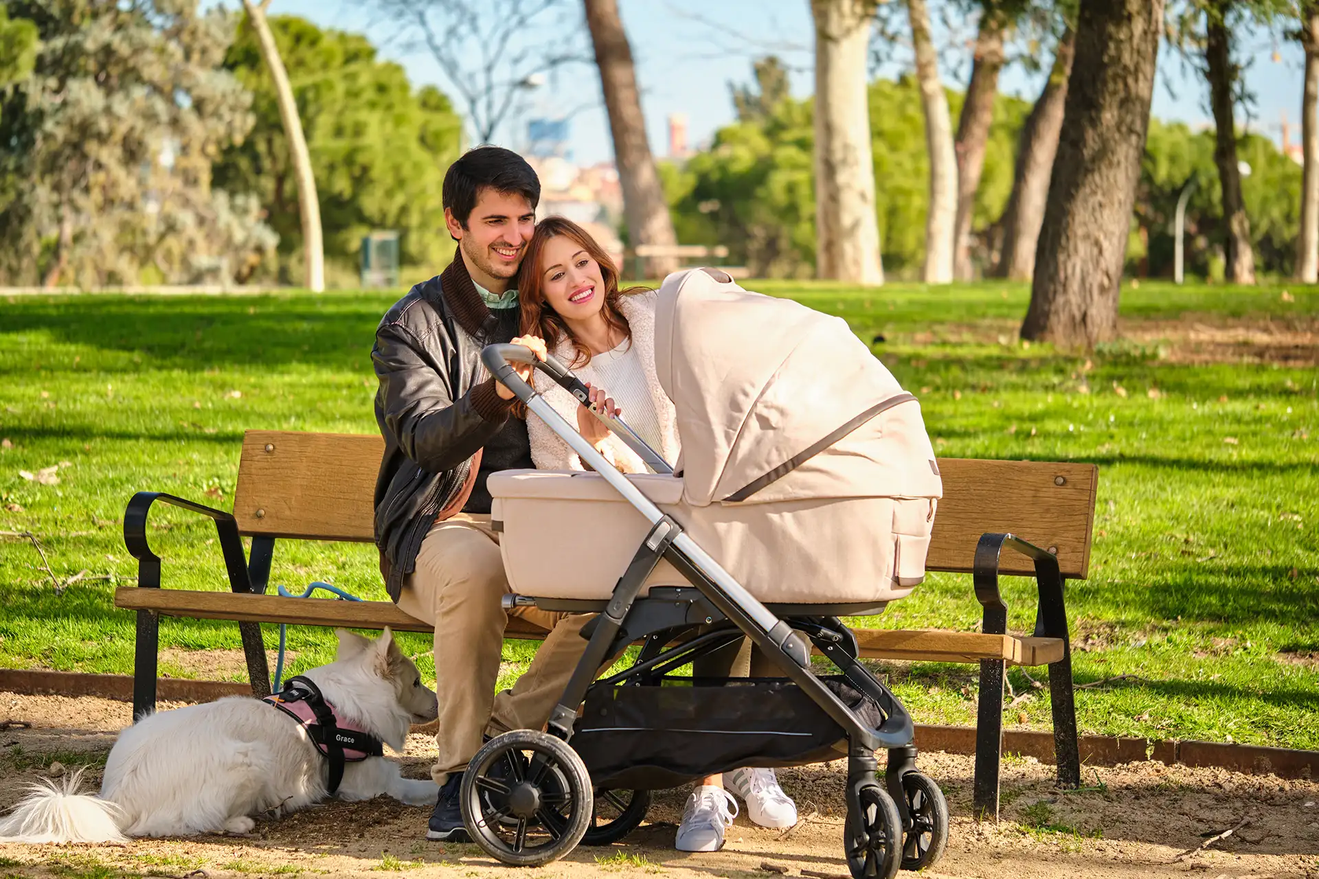 Young couple sitting on a park bench with a beige baby pram and a white dog nearby on a sunny day.