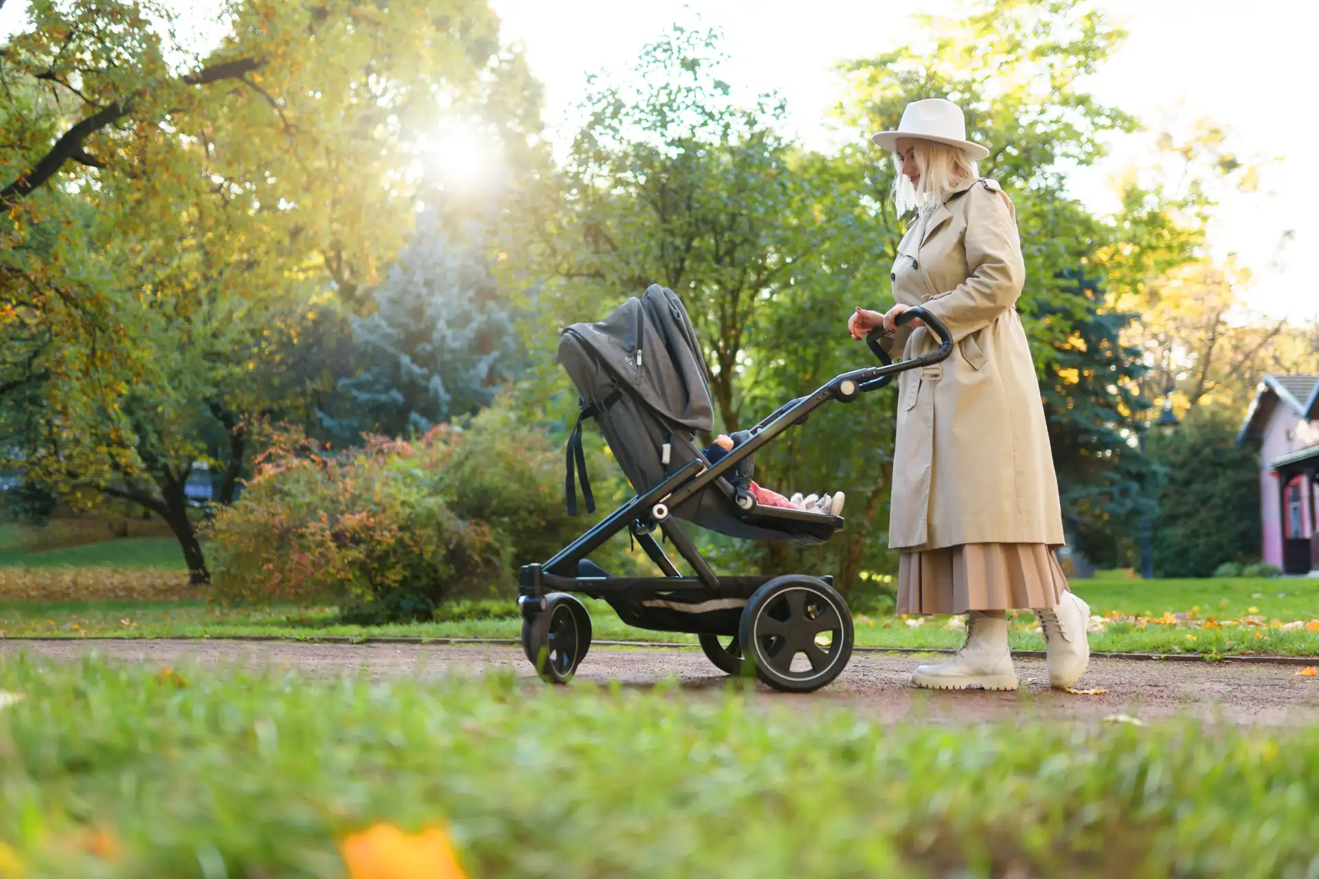Woman in beige trench coat pushing a stroller in a park during autumn - we buy your pram.